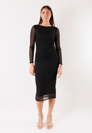 Donna in piedi rivolta in avanti, con un vestito midi nero con maniche lunghe trasparenti e orlo trasparente, abbinato a sandali con tacco chiaro.