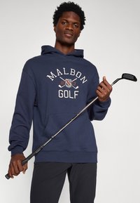 Sweat à capuche bleu marine avec "MALBON GOLF" et l'année 2017, poche avant, coupe décontractée, tenu avec un club de golf, porté avec un pantalon sombre.