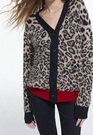Kvinde iført knappet cardigans med leopardprint, sort kant og rød ribbet kant, kombineret med sorte bukser, stående mod en ensfarvet baggrund.