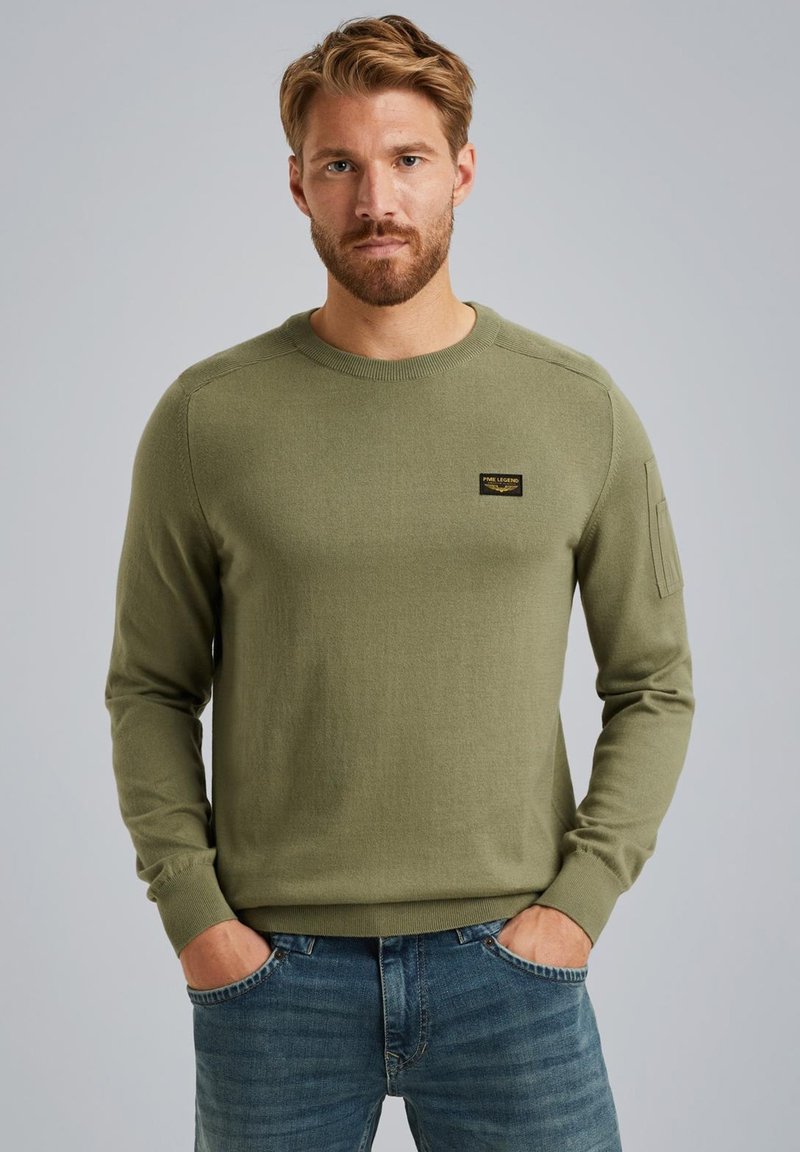 Olivno zelena pulover iz mehkega pletenega materiala z dolgimi rokavi, rebrastimi manšetami in majhno črno oznako na prsih. Uporabljeno z modrimi kavbojkami.