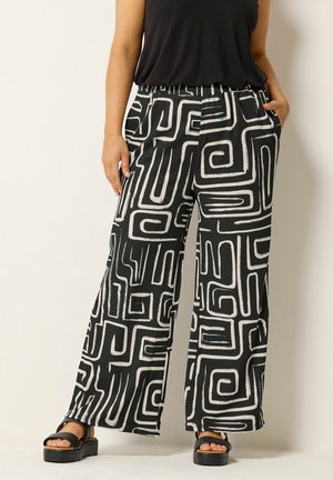 Czarno-białe spodnie o szerokich nogawkach z wyraźnym geometrycznym wzorem przypominającym labirynt, noszone z czarnym, bezrękawnym topem i czarnymi sandałami na platformie.