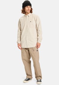 Camisa beige de botones con un bolsillo, combinada con pantalones cargo color caqui. Un gorro negro y zapatillas negras completan el look. Corte relajado y estilo casual.