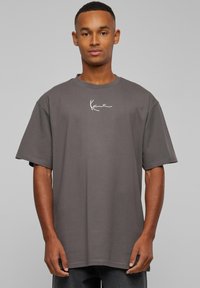 Jeune homme portant un T-shirt gris foncé ample avec un petit logo blanc centré sur la poitrine, sur un fond uni.