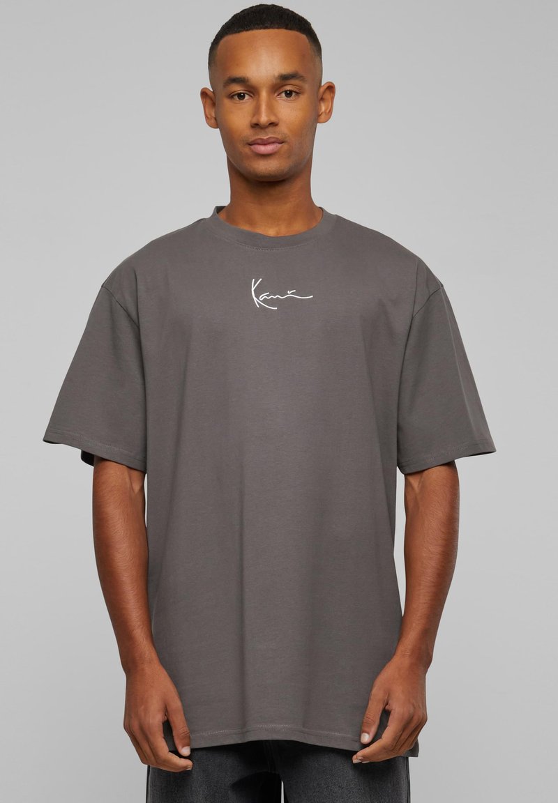 Jeune homme portant un T-shirt gris foncé ample avec un petit logo blanc centré sur la poitrine, sur un fond uni.
