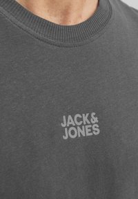 Szary sweatshirt z okrągłym dekoltem, wykonany z miękkiego materiału, z delikatnym logo "JACK & JONES" nadrukowanym w jasnoszarym kolorze na piersi.