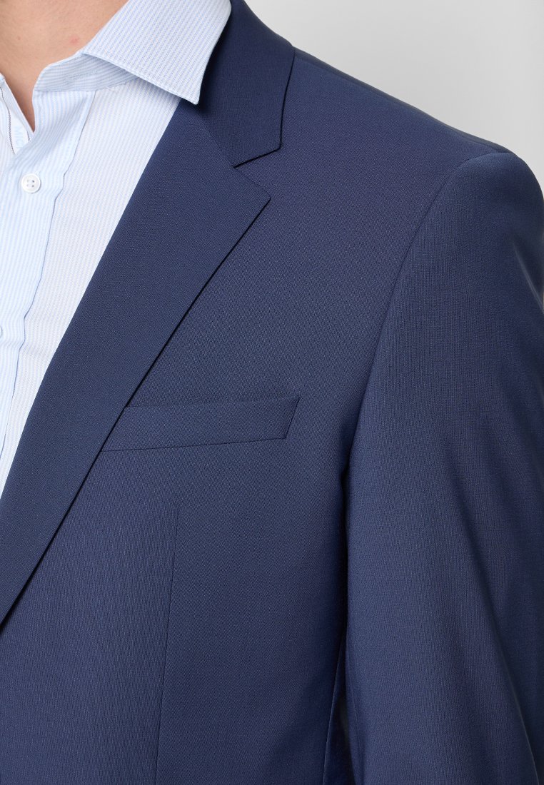 Blazer navy in tessuto texturizzato, con taschino sul petto e rever a contrasto. Indossato sopra una camicia da uomo a righe blu chiaro.