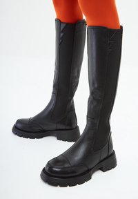Botas altas de cuero negro con suelas gruesas, llevadas sobre leggings de color naranja brillante, sobre un fondo claro.