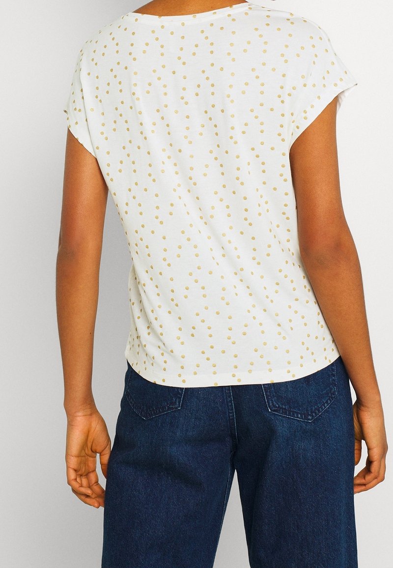 Personne portant un haut blanc à manches courtes avec des pois dorés et un jean en denim bleu foncé, vue de dos.