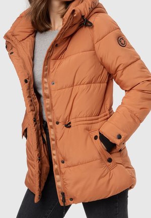 Femme portant une doudoune orange brûlé ouverte avec une capuche, un t-shirt gris en dessous et un pantalon noir, main dans la poche de la doudoune.