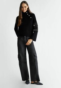 Maglione nero a coste con colletto alto, caratterizzato da dettagli con bottoni e maniche lunghe. Abbinato a pantaloni cargo neri larghi con tasche laterali e scarpe basse.