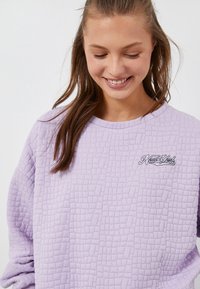Sudadera de color púrpura claro con un patrón texturizado y acolchado, y un acento bordado de "Nueva York" en el pecho. Cuello redondeado y mangas largas.