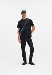 T-shirt nera e jeans scuri con una borsa a tracolla. La borsa presenta una striscia blu, una zip e una tracolla regolabile. Un cappellino nero completa il look.
