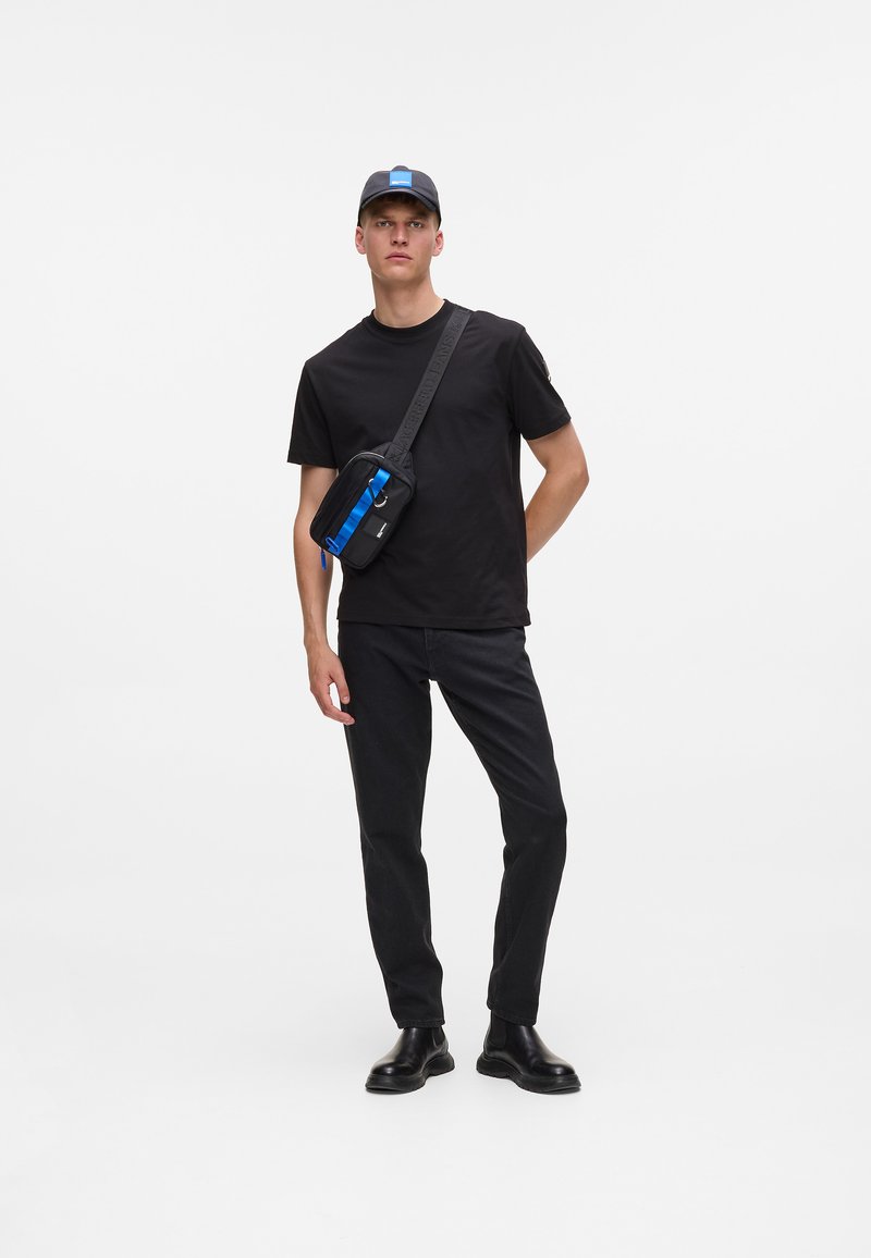 T-shirt nera e jeans scuri con una borsa a tracolla. La borsa presenta una striscia blu, una zip e una tracolla regolabile. Un cappellino nero completa il look.