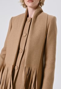 Cappotto in misto lana beige con colletto alto, cinque bottoni e lunghe frange all'orlo. La texture è liscia con una vestibilità strutturata.
