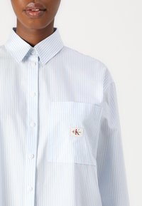 Light blue and white striped shirt with a button-down collar, chest pocket, and a logo patch. Made of smooth fabric with a relaxed fit.