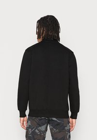 Felpa nera con collo alto, polsini e orlo a costine, realizzata in tessuto morbido, abbinata a pantaloni cargo con motivo camouflage.