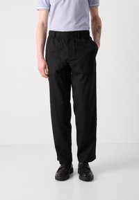 Black, loose-fitting pants with elastic waistband and side pockets, made of a textured fabric. Worn with a light purple polo shirt and black shoes.
