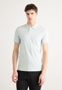 Světle modrá polo tričko vyrobené z hladkého materiálu, s klasickým límcem, tříknoflíkovým zapínáním a malou nášivkou loga na hrudi.