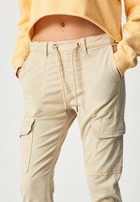 Beige cargo byxor med elastisk midjeband, dragsko, framfickor och sidofickor. Slät tyg med en lätt, hållbar textur.