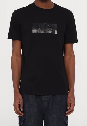 Homme portant un t-shirt noir à manches courtes et col rond avec un patch noir brillant affichant "AX" sur la poitrine, associé à un pantalon en denim bleu foncé.