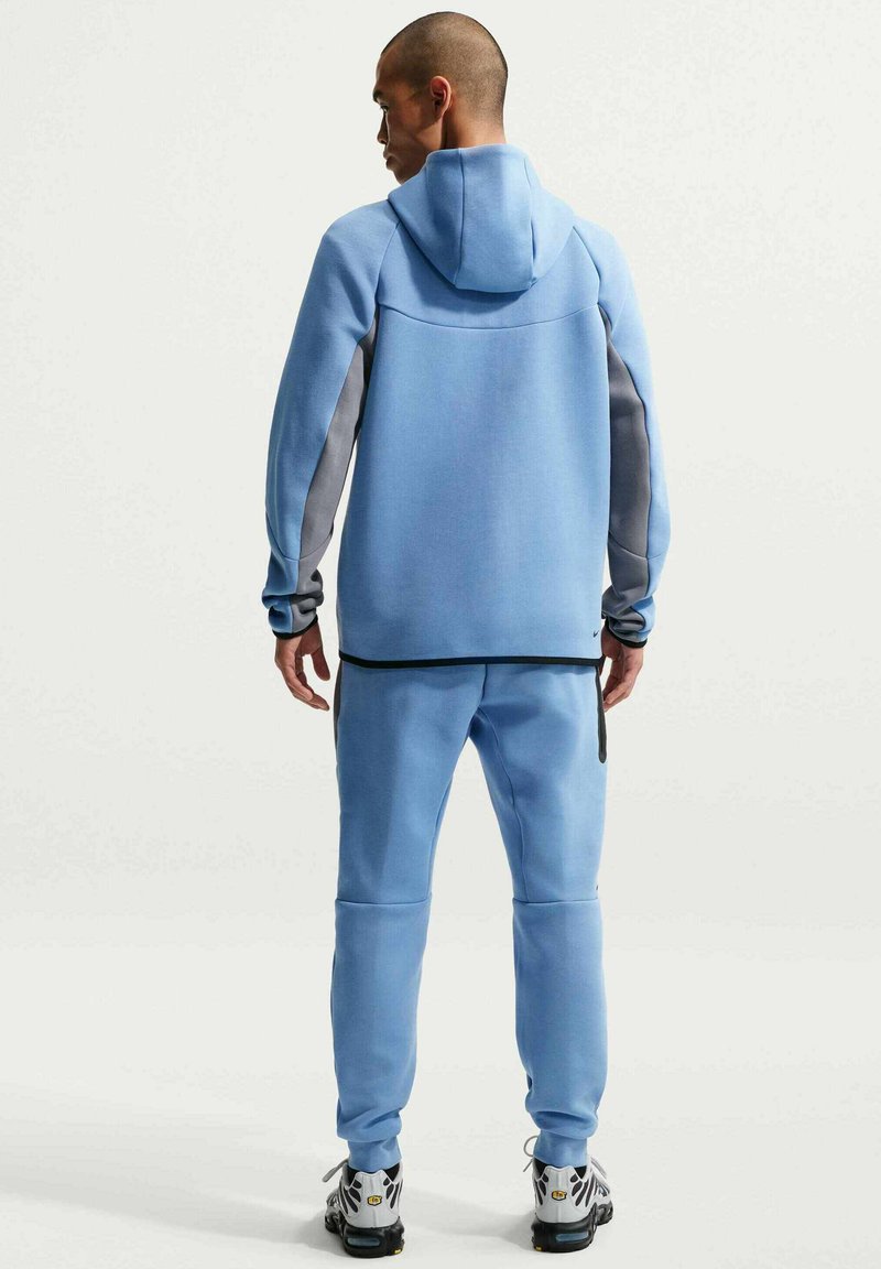 Felpa con cappuccio azzurro chiaro, dettagli grigi e vestibilità aderente, abbinata a pantaloni coordinati. Il tessuto sembra morbido con una leggera texture.