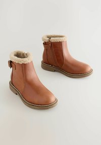 Braune Stiefeletten mit beigefarbenem Fleece-Futter, seitlichem Reißverschluss und Schnallen-Detail auf einem hellen Hintergrund.