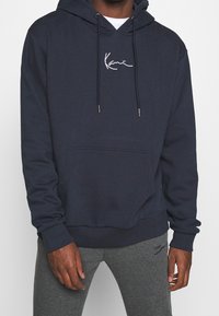 Hombre vestido con una sudadera azul marino con bolsillo frontal y logo blanco, acompañado de pantalones de chándal grises con un diseño bordado en el muslo.