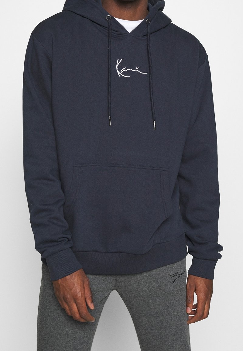 Hombre vestido con una sudadera azul marino con bolsillo frontal y logo blanco, acompañado de pantalones de chándal grises con un diseño bordado en el muslo.