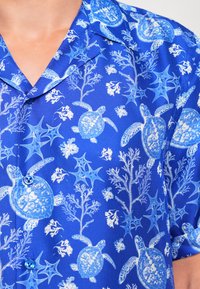 Niebieska koszula z krótkim rękawem z białymi i jasnoniebieskimi wzorami żółwi morskich oraz koralowców, z kołnierzykiem z wycięciem i niebieskimi guzikami.