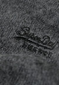 Tessuto grigio lavorato a maglia con una texture morbida, caratterizzato da un logo Superdry in rilievo di un grigio più scuro. Il design ha un aspetto sottile e testurizzato.