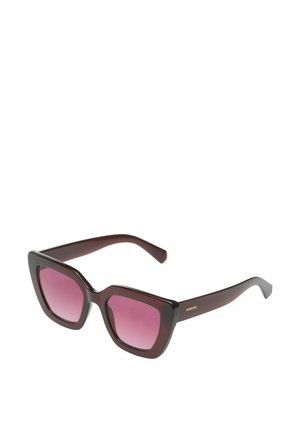 Lunettes de soleil oversize en forme d'œil de chat de couleur brun foncé avec des verres dégradés roses et le logo « KOMONO » sur la branche, vue en angle sur fond blanc.