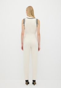 Combinaison blanche sans manches avec des accents noirs sur les épaules et la ceinture, silhouette ajustée, tissu lisse, accompagnée de talons noirs.