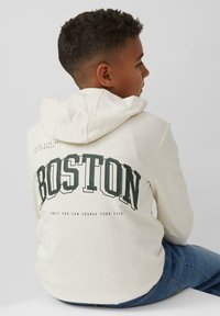 Felpa color crema con cappuccio, caratterizzata dalla scritta "BOSTON" in grassetto verde sulla schiena, con ulteriore testo sottostante. Indossata con jeans blu.