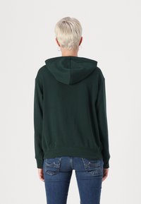 Felpa con cappuccio verde scuro, dal taglio comodo, con polsini e orlo a coste, realizzata in tessuto morbido, indossata con jeans blu, vista di spalle.