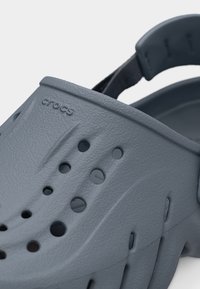 Κοντινή λήψη ενός γκρι clog Crocs που δείχνει υφή υλικού, τρύπες αερισμού και ρυθμιζόμενο πίσω λουράκι.