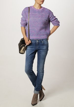 Maglione viola a coste, jeans blu slim-fit e stivali alla caviglia grigi. La modella tiene una borsa nera con tracolla.