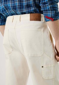 Beige cargobroek met meerdere zakken, gemaakt van katoen. Voorzien van een bruin leren patch op de tailleband en zichtbare stikseldetails.