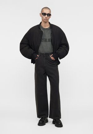 Persoon draagt een oversized zwarte bomberjacket, grijs versleten t-shirt, zwarte wijde jeans, zwarte stevige schoenen en zwarte zonnebril.
