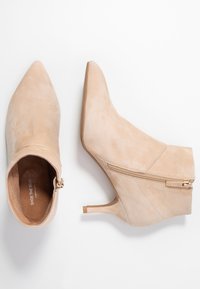 Bota de tobillo de ante beige con punta afilada, cremallera lateral y tacón delgado. Textura suave y diseño minimalista.