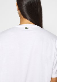 Camiseta de algodón blanca con cuello redondo, que presenta un pequeño logo de cocodrilo verde bordado en la parte de atrás, cerca del cuello.