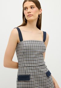 Kostkované šaty se čtvercovým výstřihem, kontrastními tmavými ramínky a denimovými akcenty na kapsách. Látka má vzor houndstooth.
