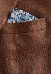 Braun strukturiertes Gewebe eines Sakko mit einem teilweise sichtbaren blau gemusterten Einstecktuch in der Brusttasche.