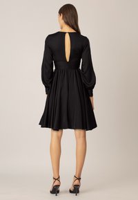 Svart klänning med utsvängt kjol, långa ärmar och en nyckelhålsdetalj i ryggen. Slät tyg med en åtsittande bodice och rynkad midja.