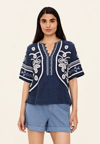 Marinblå broderad blus med vita detaljer, korta ärmar, V-ringning och dekorativa knappar. I kombination med ljusa denimshorts.