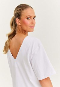 Kobieta z blond włosami związanymi w koński ogon, w białej bluzce z dekoltem w kształcie litery V, spoglądająca przez ramię na tle jasnym.