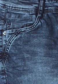Jeans en denim bleu avec une texture lisse, comportant une poche avant et des coutures contrastantes. Le tissu a un aspect usé et légèrement décoloré.