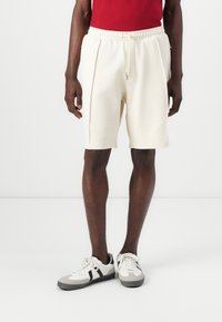 Shorts de couleur crème avec une taille élastique, détails de cordon de serrage et poches latérales. Portés avec un haut rouge et des baskets avec des accents gris.