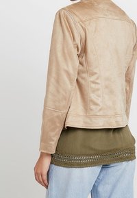 Béžová bunda z umělého semiše s přiléhavým střihem, viditelným šitím a vykrojenými zády. Nošeno přes olivově zelený top s texturovaným lemem.