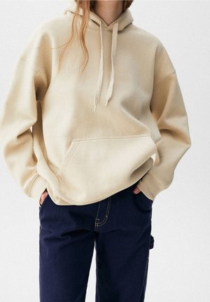 Person wearing a beige hoodie with front pocket and drawstrings, paired with dark blue pants, hands in pockets, against a plain light background.