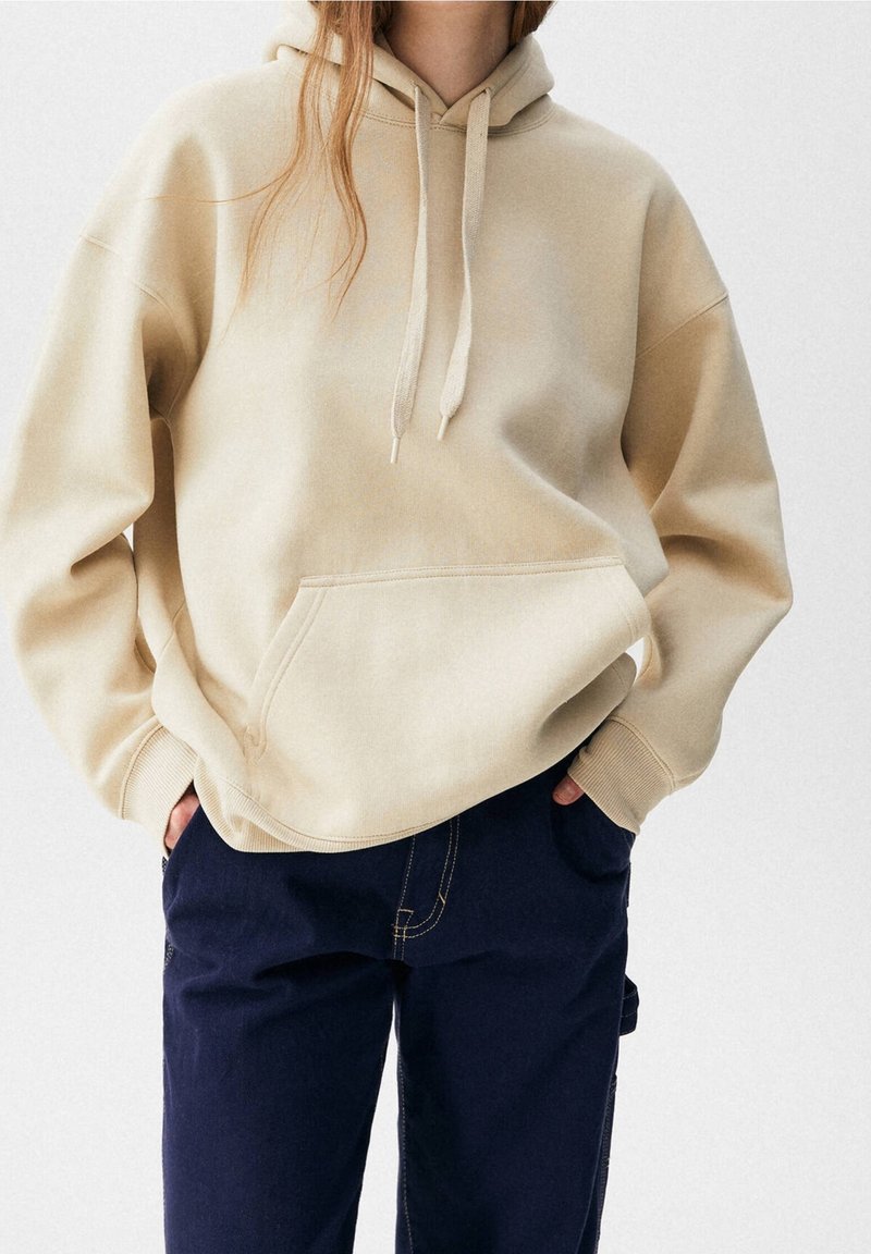 Personne portant un sweat à capuche beige avec poche avant et cordons, associé à un pantalon bleu foncé, mains dans les poches, sur un fond clair uni.